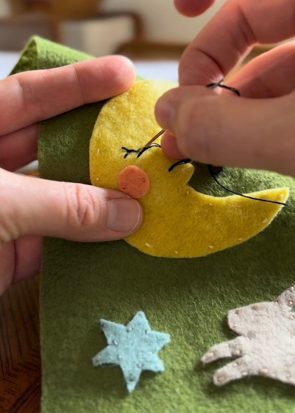 Hand sewing a yellow felt moon onto green felt fabric with other decorative elements.